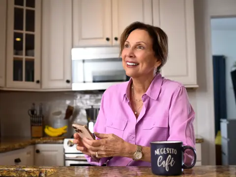 A smiling woman in a pink shirt sits at a kitchen counter, holding a smartphone and looking at it.