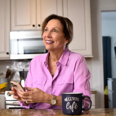 A smiling woman in a pink shirt sits at a kitchen counter, holding a smartphone and looking at it.