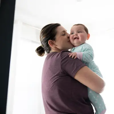 A mother holding her baby and kissing her cheek in a bedroom with a lamp.