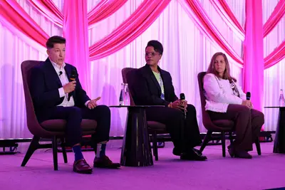 Three panelists seated on stage during a conference discussion, with one speaker addressing the audience with a microphone.