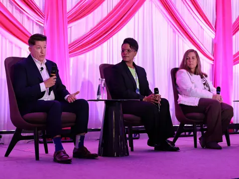 Three panelists seated on stage during a conference discussion, with one speaker addressing the audience with a microphone.