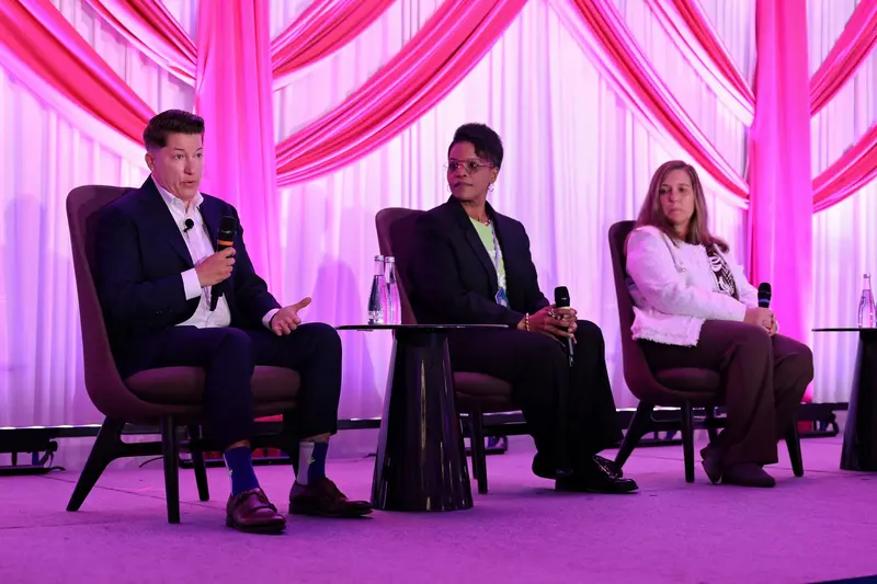 Three panelists seated on stage during a conference discussion, with one speaker addressing the audience with a microphone.