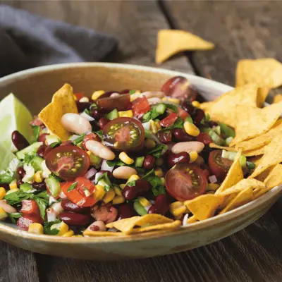 Colorful bean and corn salad with cherry tomatoes, lime wedges, cilantro, and tortilla chips in a rustic bowl.