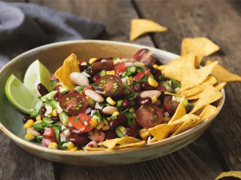 Colorful bean and corn salad with cherry tomatoes, lime wedges, cilantro, and tortilla chips in a rustic bowl.