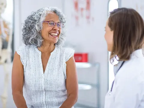 Senior woman with gray hair and glasses smiling warmly while speaking with a female doctor in a white coat in a bright medical clinic