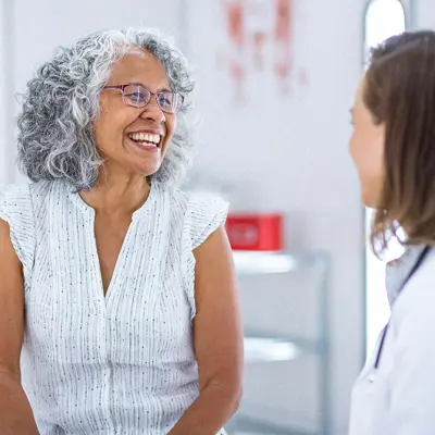 Senior woman with gray hair and glasses smiling warmly while speaking with a female doctor in a white coat in a bright medical clinic