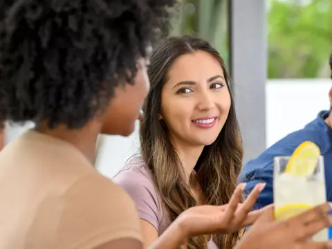 A smiling woman engages in conversation with another woman, holding a glass of lemonade.