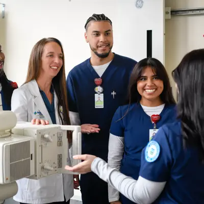 Five radiography students in navy scrubs and a white lab coat gathered around an X-ray machine in a clinical training room, smiling and engaging with each other during a hands-on learning session.