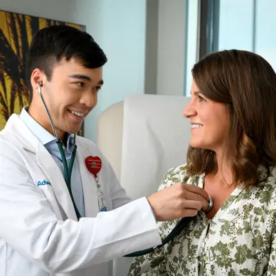 A smiling doctor in a white coat uses a stethoscope on a smiling woman in a green floral shirt.
