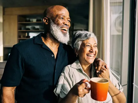 An older couple smiling and embracing while standing near a window in their home, with the woman holding an orange coffee mug