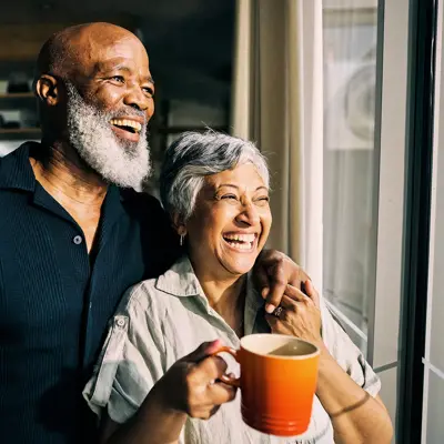 An older couple smiling and embracing while standing near a window in their home, with the woman holding an orange coffee mug