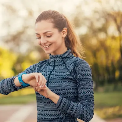 A smiling woman in athletic wear checking her fitness watch outdoors in a park setting with trees in the background