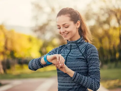A smiling woman in athletic wear checking her fitness watch outdoors in a park setting with trees in the background