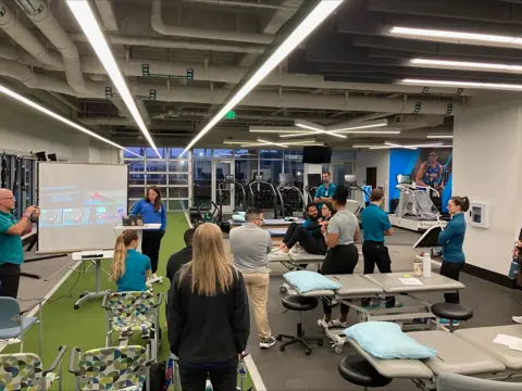 Group listens to a presenter with slides projected in a rehab gym setting.