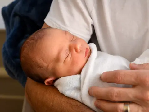 A person holds a sleeping newborn wrapped in a white blanket, conveying warmth and care.