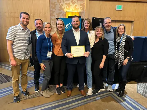 Group poses with a man holding a framed award certificate at an event.