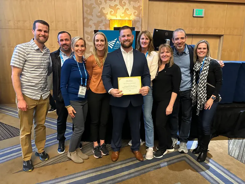 Group poses with a man holding a framed award certificate at an event.