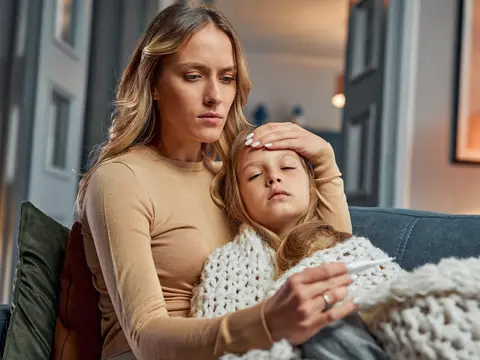 Woman holds a resting child on a couch, touching the child's forehead while holding a thermometer.
