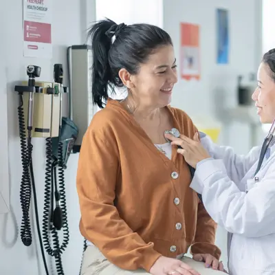A clinician uses a stethoscope on a smiling woman‚Äôs chest in a medical exam room.