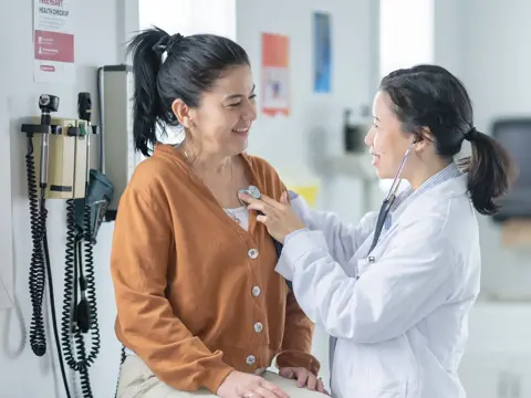 A clinician uses a stethoscope on a smiling woman’s chest in a medical exam room.