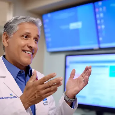 A smiling doctor in a white lab coat stands in front of multiple monitors.