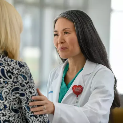 A doctor in a white coat and green shirt with a red heart badge talks to a patient.