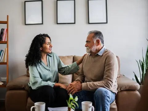 An older man sits on a couch, being comforted by a female therapist.