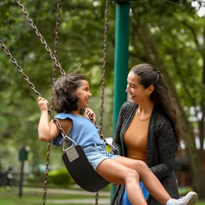 A smiling woman pushes a young girl on a swing in a park.