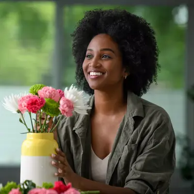 A smiling woman holds a vase of pink and white flowers, conveying warmth and positivity.