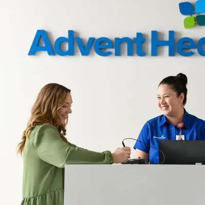 A smiling woman in a green blouse interacts with a smiling woman in a blue AdventHealth uniform at a reception desk.