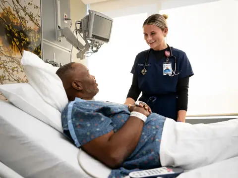 A smiling nurse stands beside a patient lying in a hospital bed, creating a warm and engaging atmosphere.