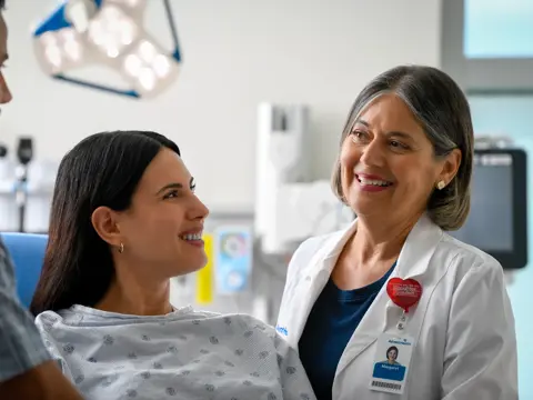 A smiling patient in a hospital gown talks with a doctor in a white coat.