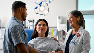 A smiling doctor and a patient in a hospital gown hold hands with a family member in a hospital room.