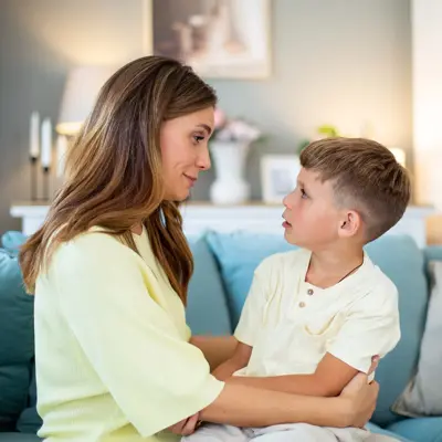 Adult and young child sit on a sofa, facing each other and holding arms in a softly lit living room.