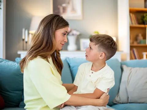 Adult and young child sit on a sofa, facing each other and holding arms in a softly lit living room.