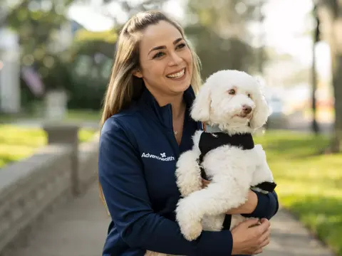 A smiling woman in a blue AdventHealth jacket holds a white dog with a harness.