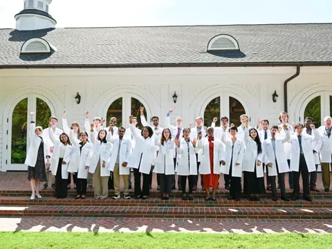 Group wearing white coats standing on brick steps in front of a white building, raising arms.