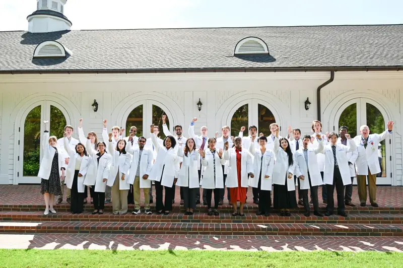 Group wearing white coats standing on brick steps in front of a white building, raising arms.