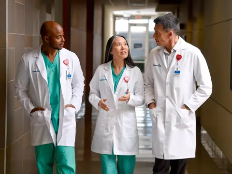 Three medical professionals in white coats and green scrubs walking down a hospital corridor.