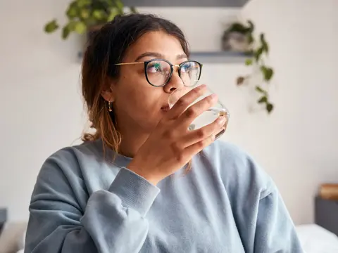 Woman with glasses drinking a glass of water while seated in a bright home interior with plants in the background