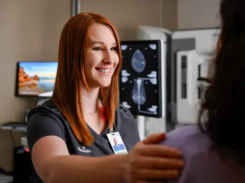 A smiling healthcare professional in a black uniform with a badge stands next to a patient in a medical setting.