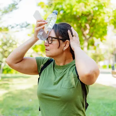 Woman in green shirt and glasses cooling herself with a water bottle pressed against her forehead while outdoors in summer heat