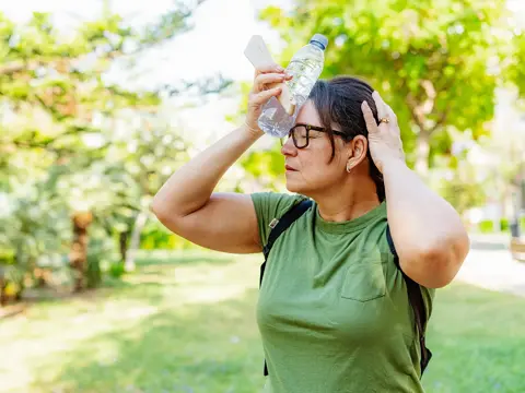 Woman in green shirt and glasses cooling herself with a water bottle pressed against her forehead while outdoors in summer heat