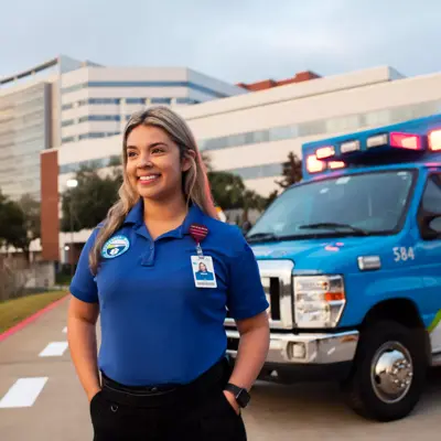 A smiling woman in a blue uniform stands next to an ambulance in front of a hospital.