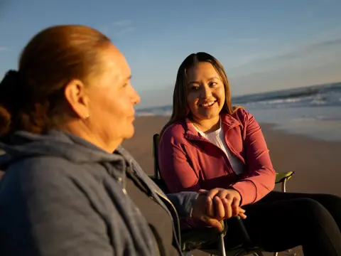 Two women sitting on the beach, one in a chair, smiling and talking.