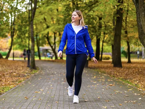 A woman in a bright blue jacket walks along a tree-lined park path surrounded by autumn foliage.