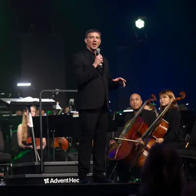 Richard Hickam speaks into a microphone onstage with seated orchestra musicians behind him.