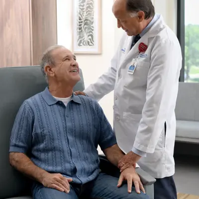 A smiling patient sits in a chair as a doctor stands beside him, placing a hand on his shoulder.