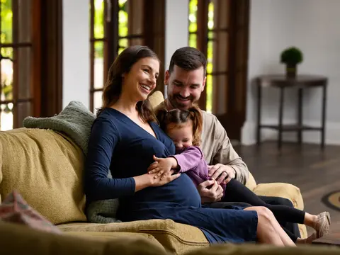 Pregnant woman in blue dress sitting on yellow couch with partner and young daughter in a bright home living room