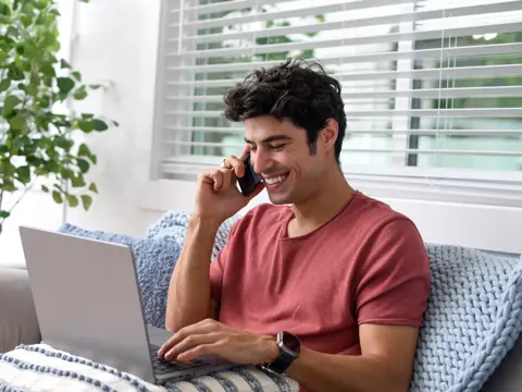 Man sitting on a couch, using a laptop and talking on the phone.
